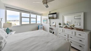 A bedroom with a large bed, a ceiling fan, a TV, a dresser, and a window with a view of the beach.