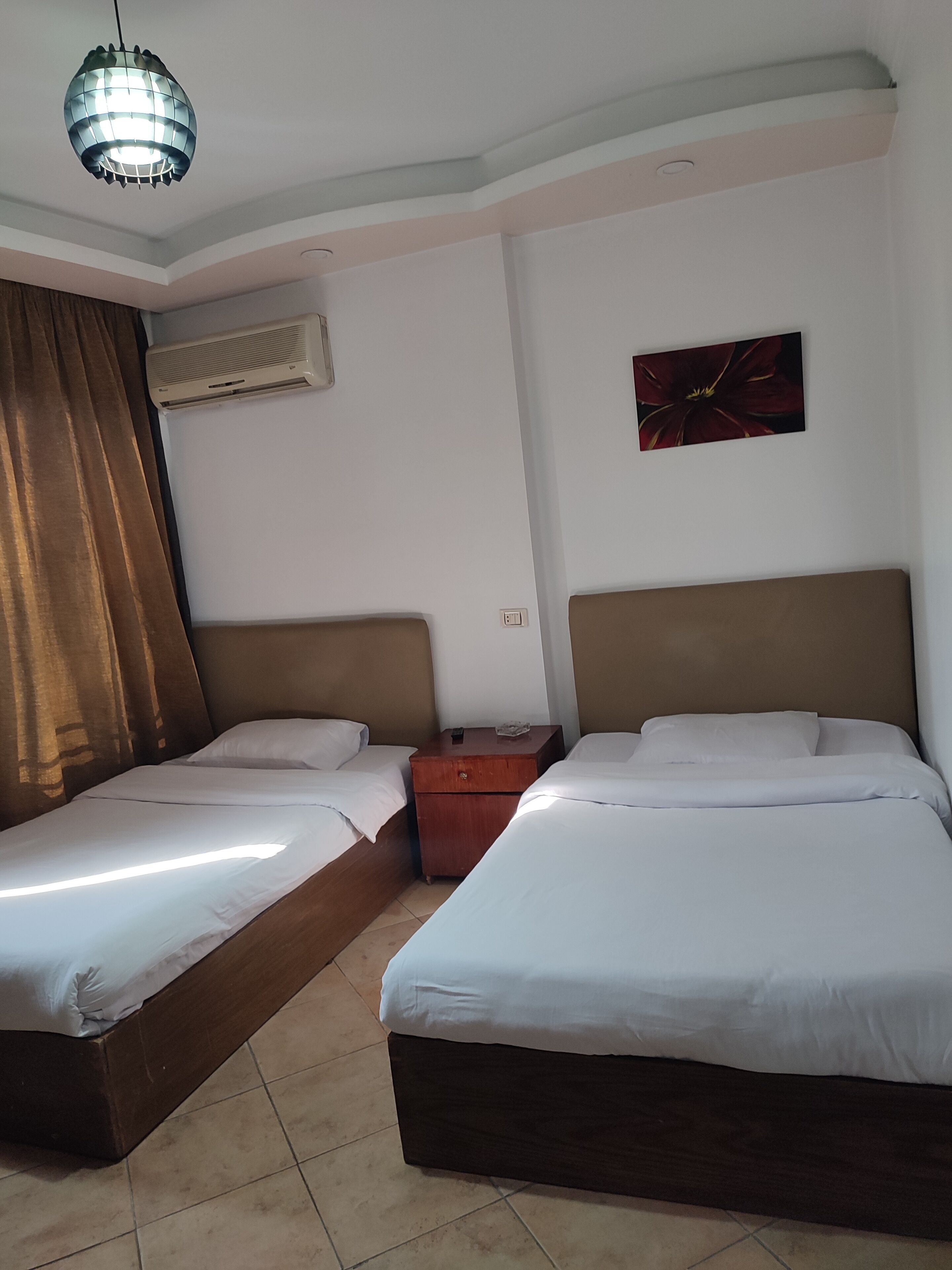 Une chambre d’hôtel avec deux lits simples, une table de chevet en bois, un climatiseur fixé au mur, un luminaire au plafond et une œuvre d’art encadrée accrochée au mur.