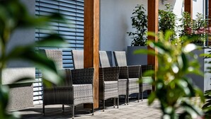 Un patio con mobiliario de mimbre, suelo de baldosas y un edificio con vigas de madera y una ventana con persianas.