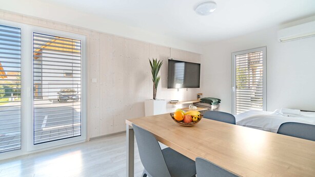 Habitación de hotel moderna con una mesa de comedor de madera, sillas grises, un televisor de pantalla plana en la pared y un amplio ventanal con persianas.