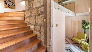 Un escalier en bois mène à une pièce dont le mur est en pierre, et qui contient une chaise et une plante.
