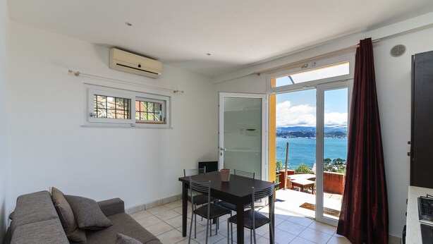 Une pièce comprenant une table à manger, des chaises, un canapé et une porte-fenêtre coulissante donnant sur un balcon avec vue sur la mer.