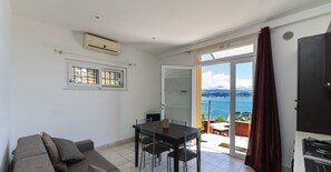 Quarto com mesa de jantar, cadeiras, sofá e porta de vidro deslizante para uma varanda com vista para o mar.