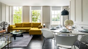 Ein helles Wohnzimmer mit einem gelben Sofa, einem Glastisch und einem Essbereich mit einem weißen Tisch, gedeckt für vier Personen.