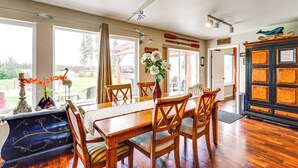 A dining area with a wooden table, chairs, and a vase with flowers. There is a window with curtains, a door, and a cabinet with a decorative item on top. - Soldotna Home w/ Yard, Walk to Kenai River! (Soldotna)