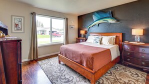 A bedroom with a large bed, a wooden dresser, a nightstand, a wall-mounted lamp, a window with curtains, and a decorative fish mural. - Soldotna Home w/ Yard, Walk to Kenai River! (Soldotna)