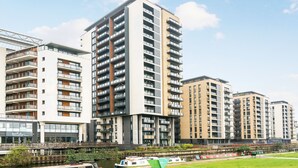 A high-rise residential building with multiple balconies, a river in the foreground, and a small bridge. - Neumann (London)