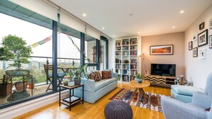 A spacious living room with a large window, a sofa, a TV, a bookshelf, and a potted plant. - Neumann (London)
