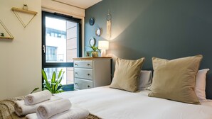 A neatly made bed with rolled towels, a bedside lamp, and a window with a view of buildings. - Neumann (London)