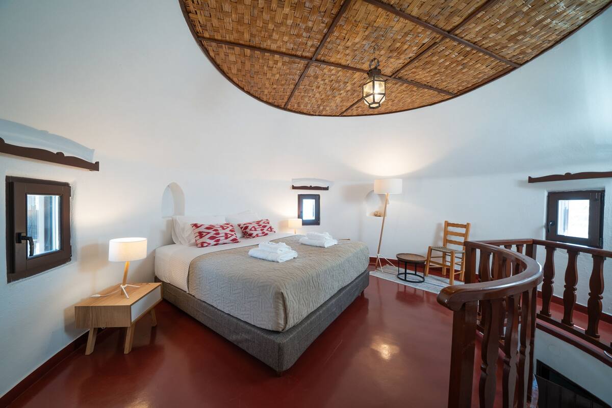 A bedroom with a large bed, a small table, a chair, a lamp, and a balcony railing.