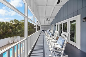 A covered outdoor area with white rocking chairs, a pool, and a view of palm trees.