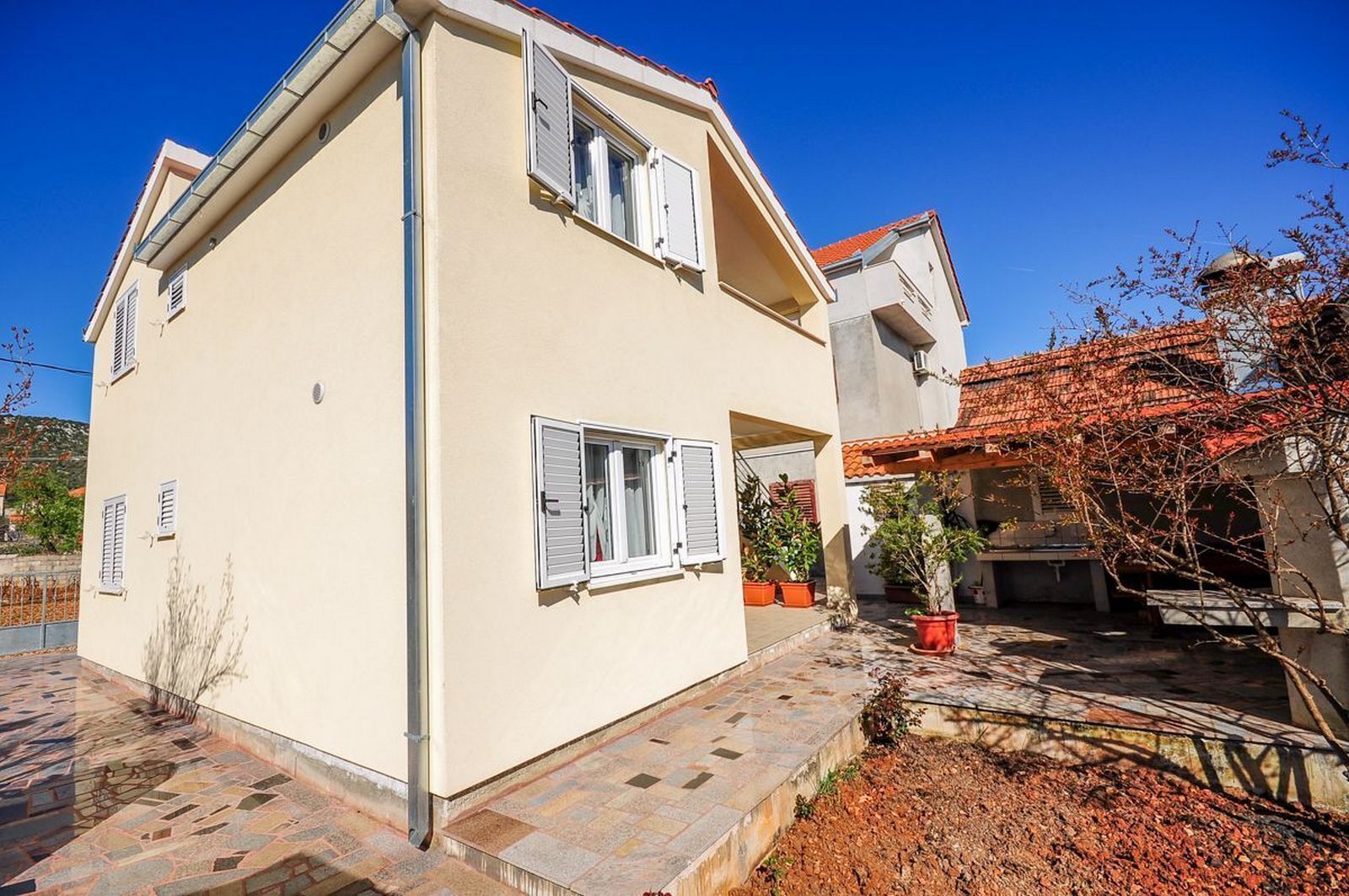 A two-story building with a beige exterior, multiple windows with shutters, and a tiled patio area with potted plants.