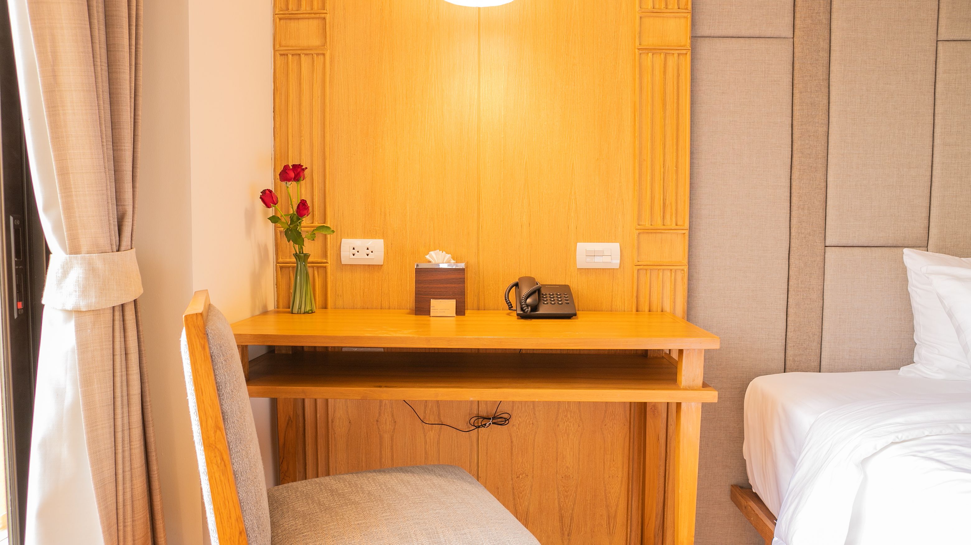 Una habitación de hotel con un escritorio de madera, un teléfono, un florero con flores y una mesita de noche.