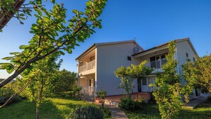 Ein zweigeschossiges Haus mit Balkon im oberen Bereich, umgeben von viel Grün und blauem Himmel.
