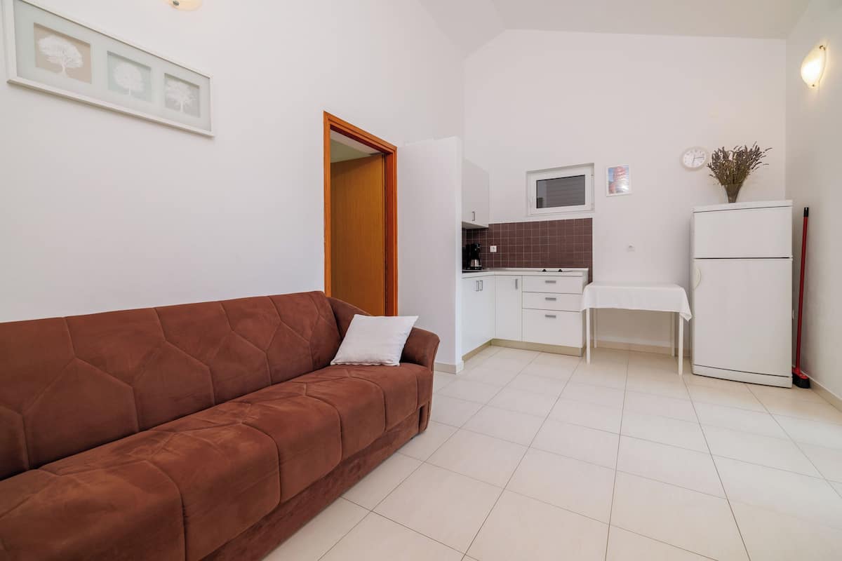 Una sala de estar con un sofá marrón y una mesa blanca. Hay una zona de cocina con refrigerador, microondas y alacenas.