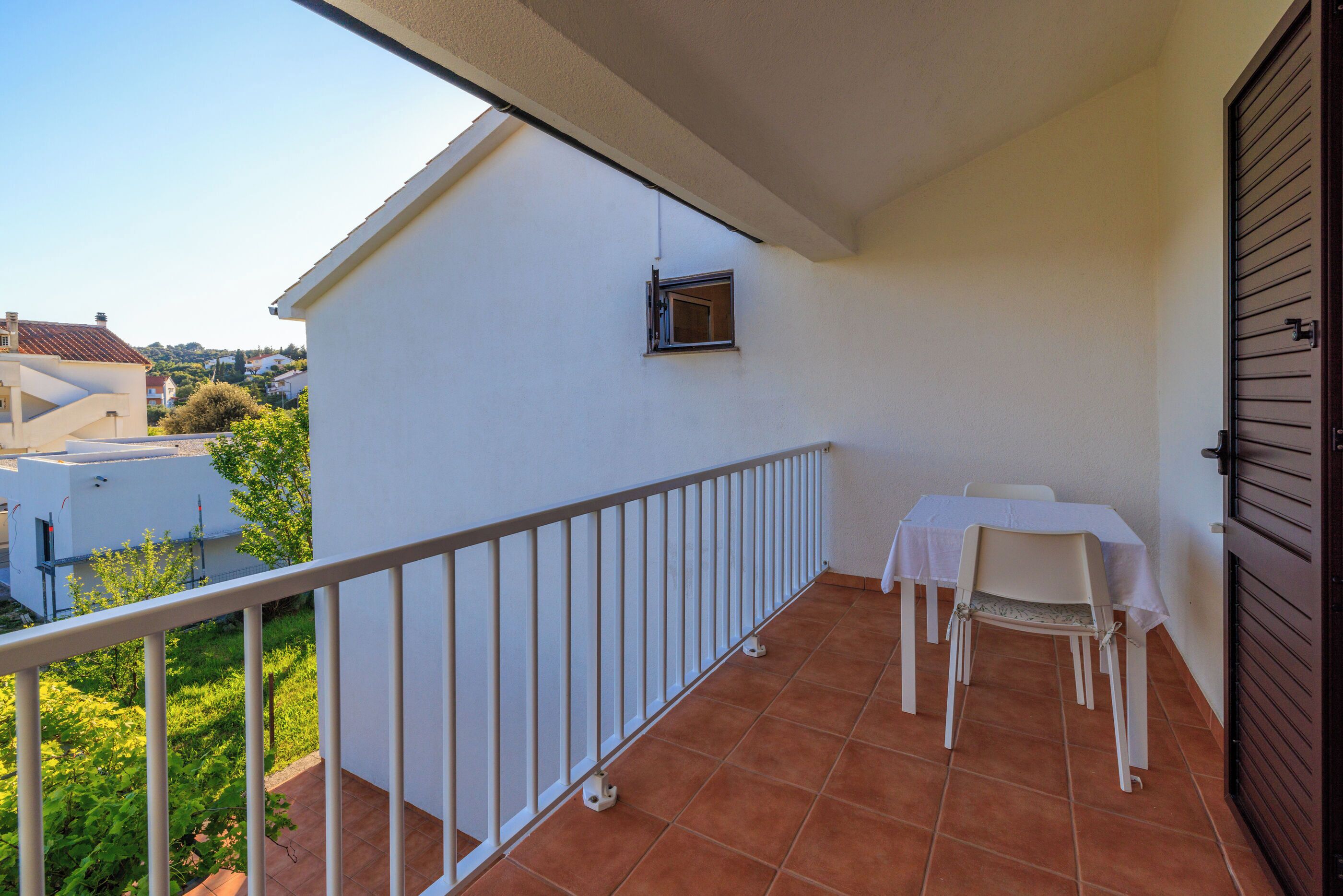 A balcony with a white table and chairs, a small window, and a view of the neighborhood.