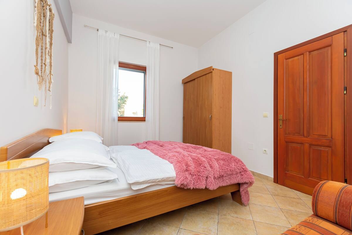 A bedroom with a wooden bed, a pink blanket, a wooden wardrobe, and a window with curtains.