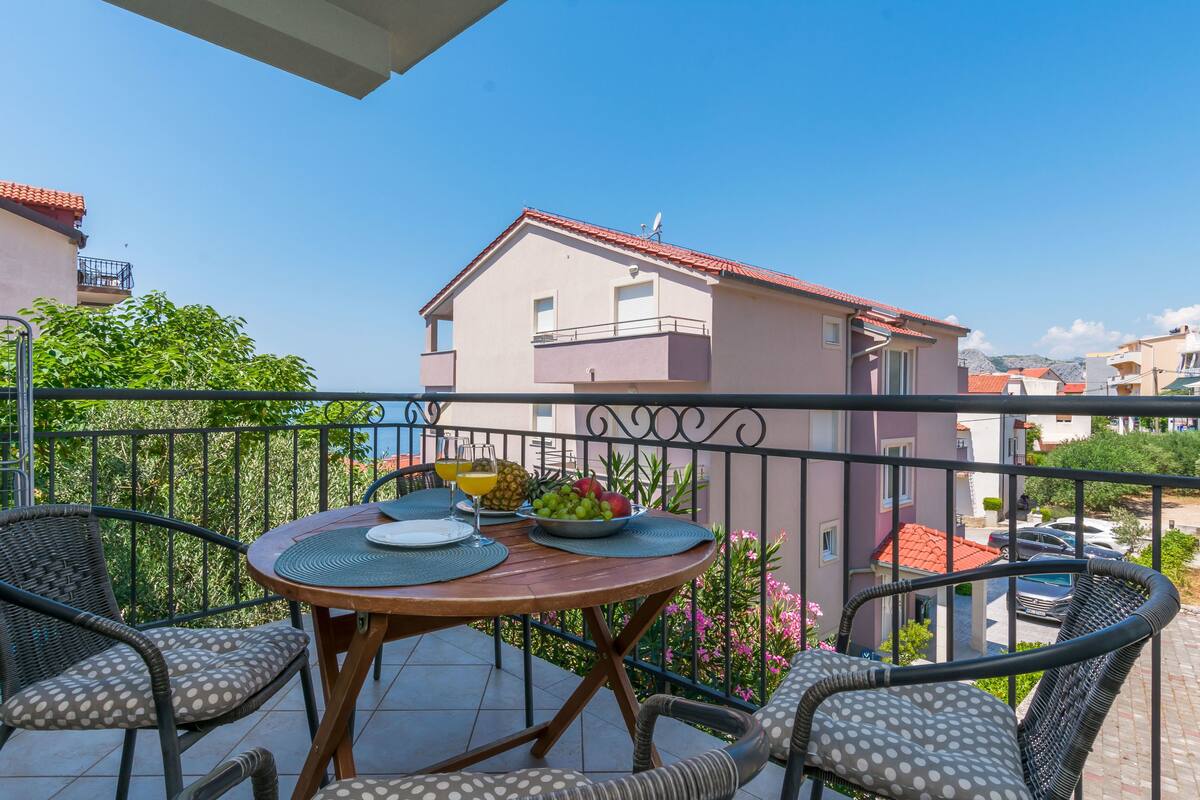 A balcony with a table set for two, surrounded by wicker chairs, overlooking a residential area with houses and a clear sky.