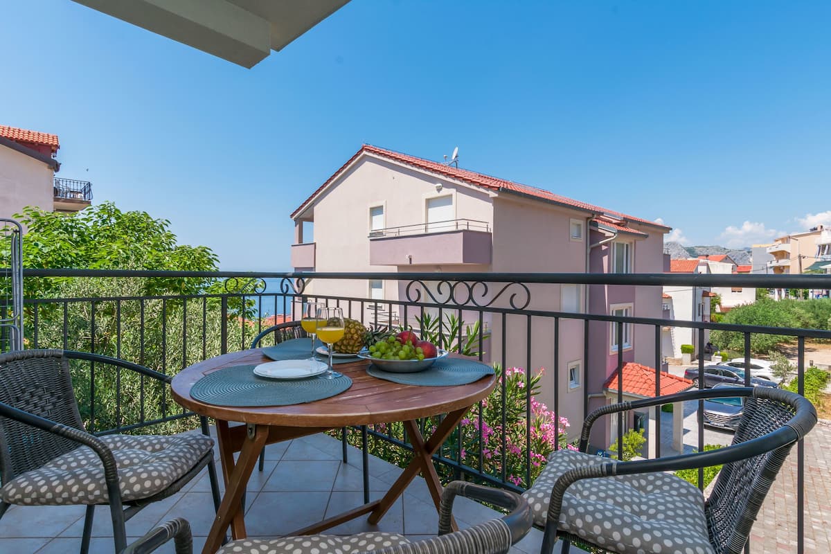 A balcony with a table set for two, surrounded by wicker chairs, overlooking a residential area with houses and a clear sky.