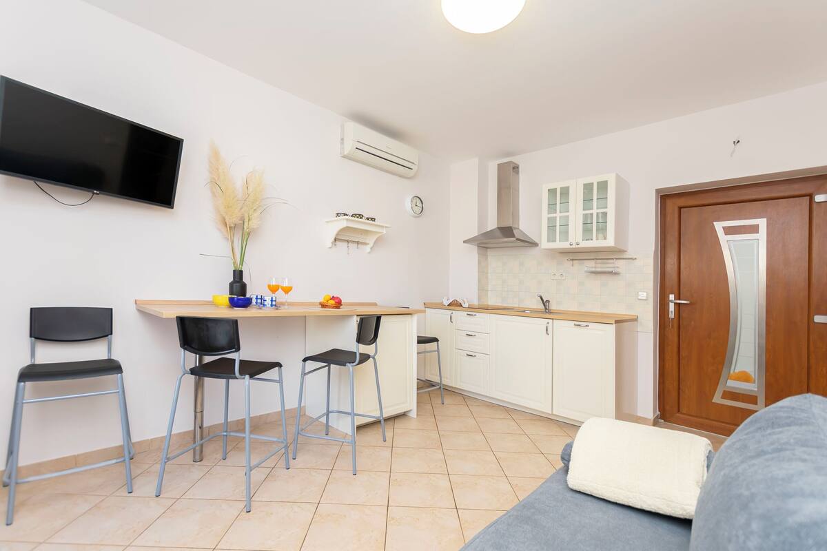 A compact kitchen with a flat-screen TV, a dining area with chairs, a small table with fruits, and a sofa.