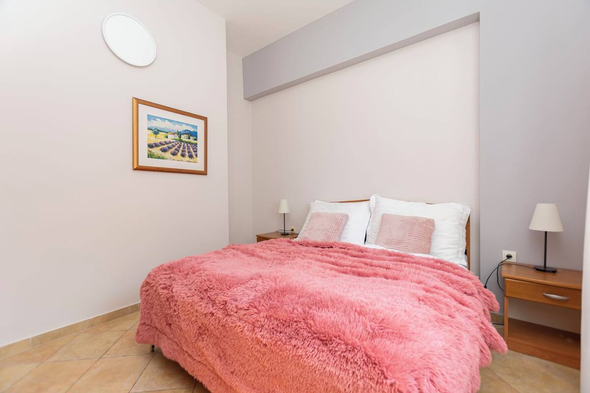 A bedroom with a pink fluffy bedspread, a nightstand with a lamp, a framed picture on the wall, and a wooden desk with a lamp.