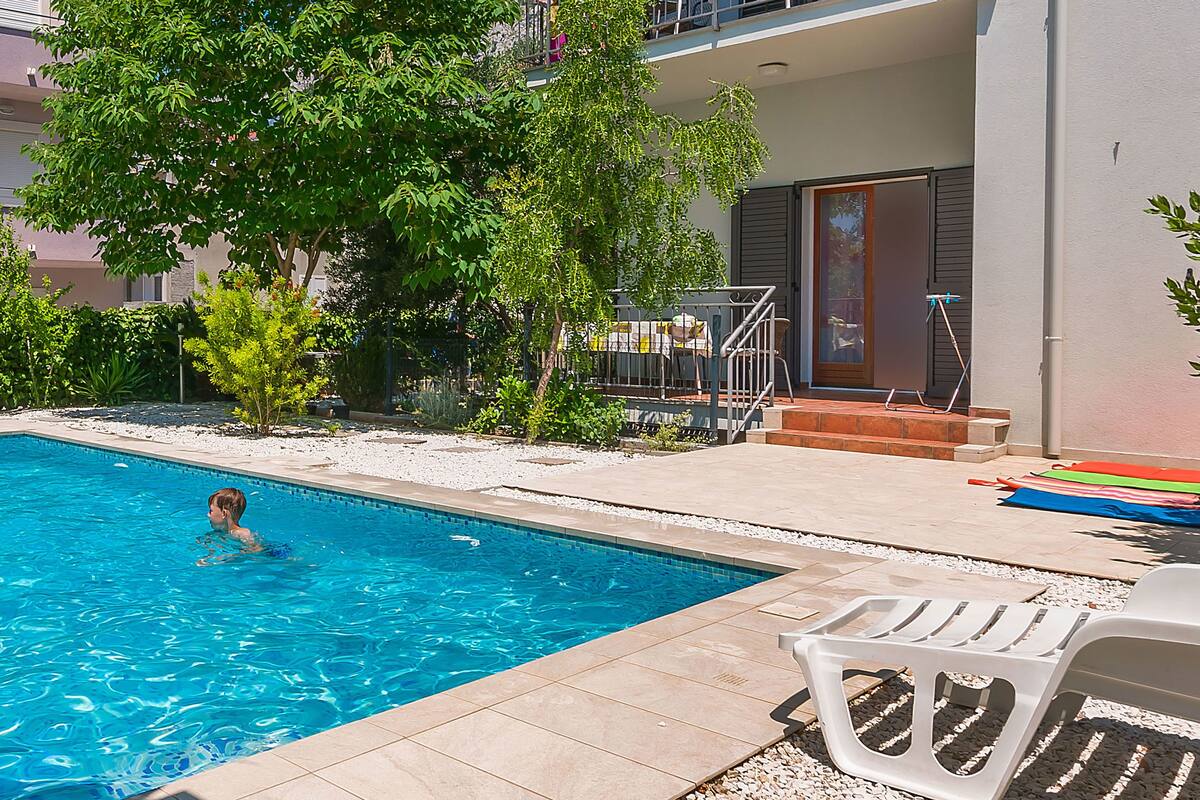 A swimming pool with a child swimming, surrounded by a patio area with chairs and a building in the background.