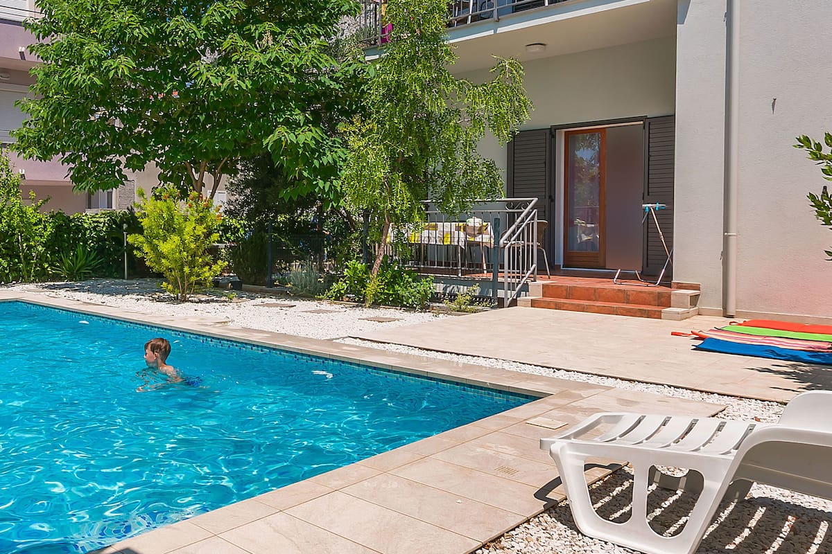 A swimming pool with a child swimming, surrounded by a patio area with chairs and a building in the background.