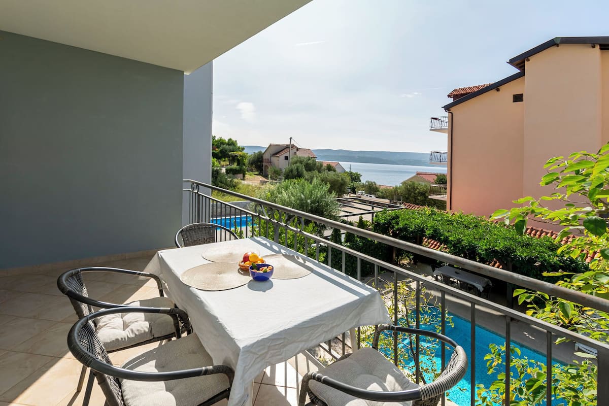 A balcony with a table and chairs, overlooking a swimming pool and a view of the sea and houses.