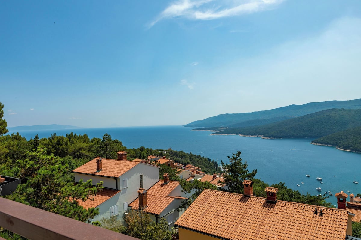 A coastal village with terracotta-roofed houses, a clear blue sea, and boats anchored nearby.