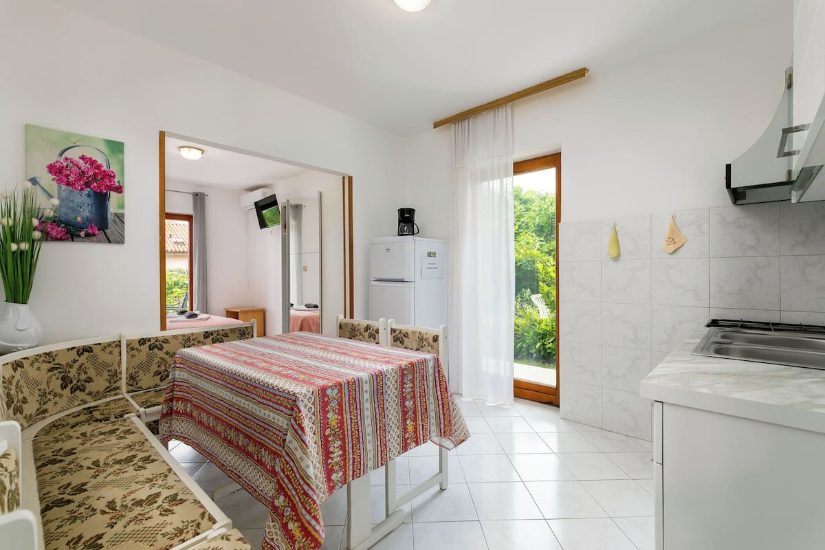 A compact room with a dining table, a sofa, a kitchen area, and a view of greenery through a window.
