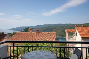 A balcony with a table and chairs, overlooking a coastal landscape with buildings and hills.