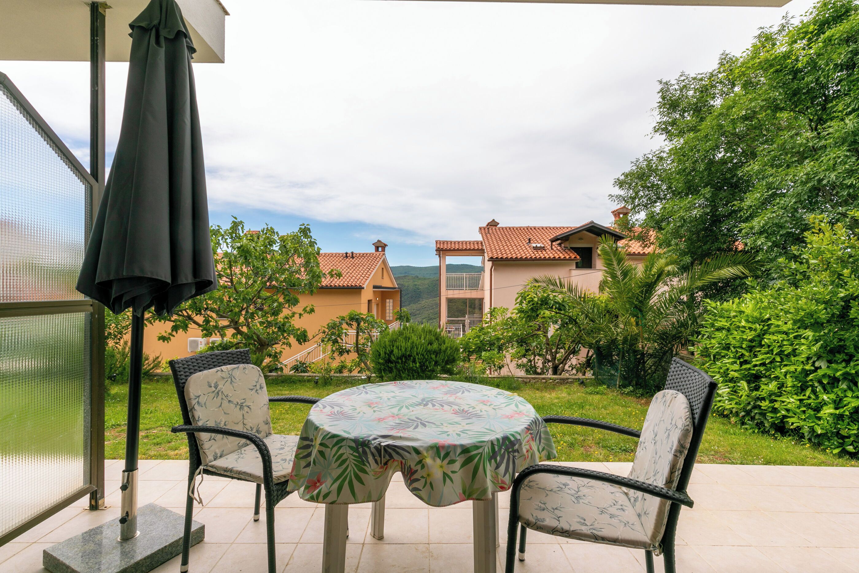 A patio with a round table and two chairs, a covered umbrella, and a view of buildings and greenery.