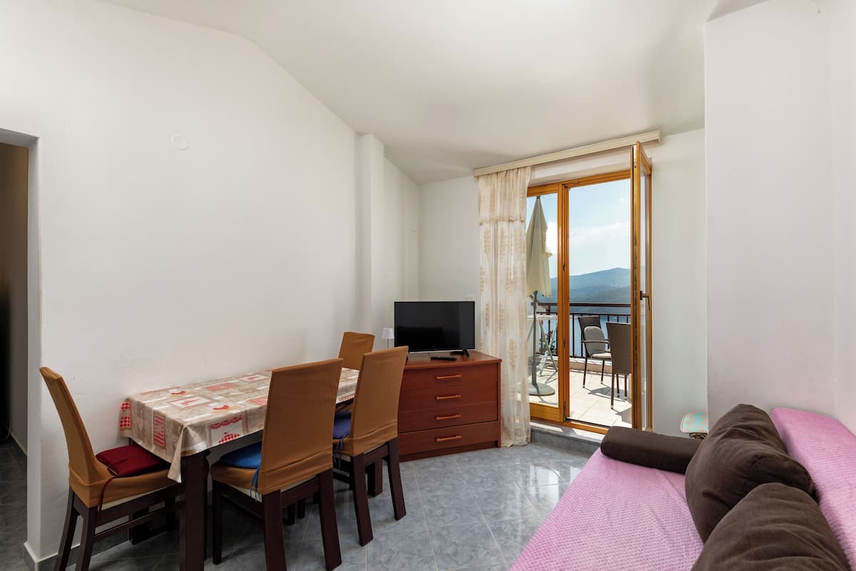 A bedroom with a bed, a TV, a dining table with chairs, and a balcony with a view.