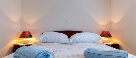 A neatly made bed with a checkered bedspread, two white pillows, two red lamps on nightstands, and two rolled blue towels.