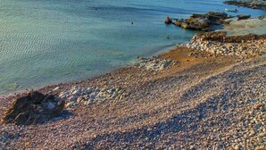 Una playa de piedras con agua tranquila, barcos y afloramientos rocosos.