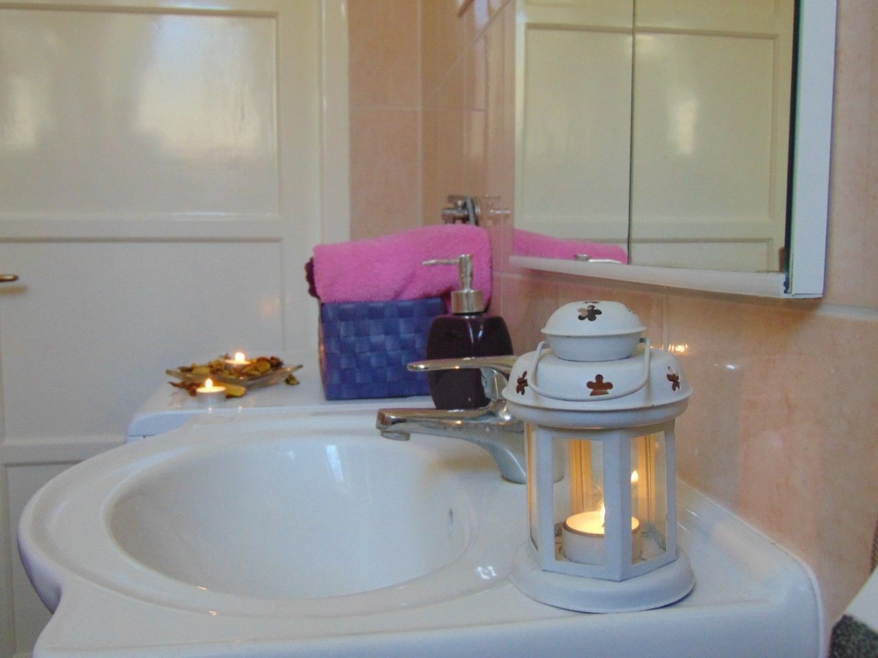Un lavamanos de baño con un lavabo blanco, una toalla rosa, un dispensador de jabón y un farol blanco con una vela en su interior.