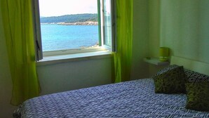 Un dormitorio con cama, ventana con cortinas, mesita de noche con lámpara y vista al mar.