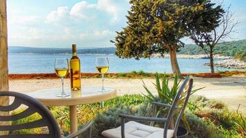 A table with a bottle and two glasses of wine, a chair, and a view of the sea and hills.