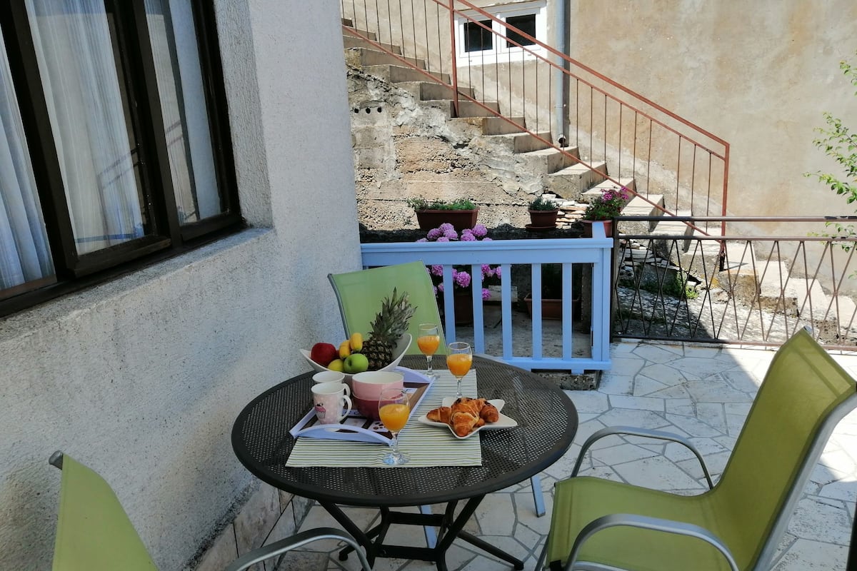 Une terrasse avec une table dressée pour le petit-déjeuner, comprenant des fruits, des croissants et du jus d’orange.