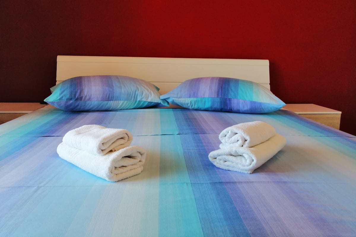Un lit bien fait, recouvert d’une couverture aux couleurs dégradées, allant du bleu au violet, avec deux oreillers et deux serviettes pliées.