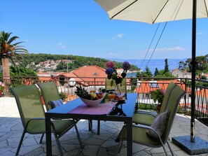 A balcony with a table set for two, featuring a bowl of fruit, a glass of juice, and a cup. The balcony overlooks a scenic view of a coastal town with red-roofed buildings and a clear blue sky. - Apartments Amazing View (Jelsa)