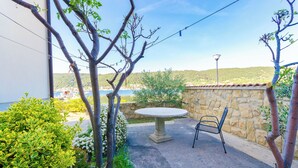 A patio with a stone wall, a round table, and a chair, overlooking a body of water and hills. - Apartments Darko (Supetarska Draga)