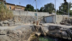A rocky pool area with steps leading into the water, surrounded by a stone wall and a building with a satellite dish. - Apartments Romina (Rijeka)