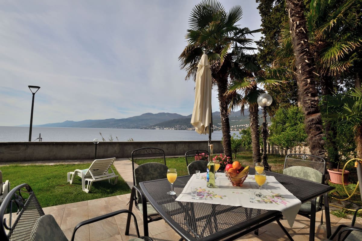 An outdoor dining area with a table set for two, overlooking a scenic view of the sea and mountains.