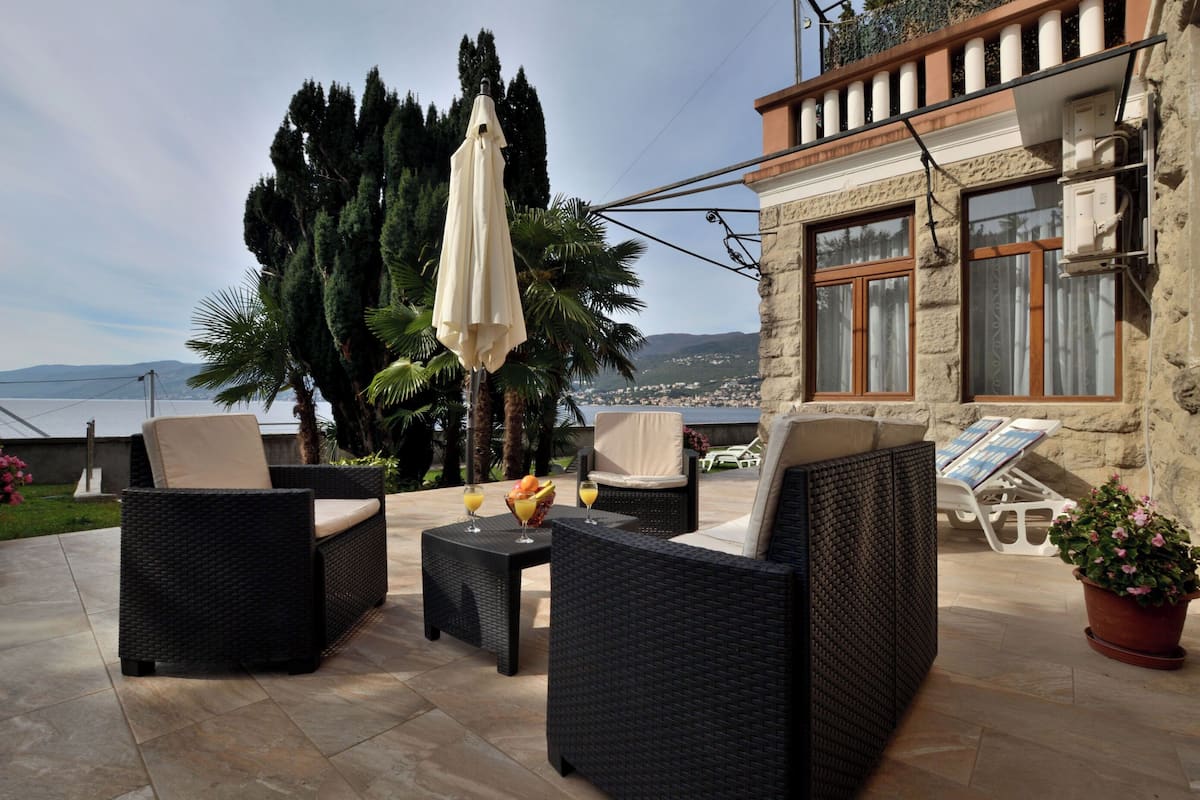 An outdoor seating area with wicker furniture, a table with fruit and drinks, and a view of the sea and hills.