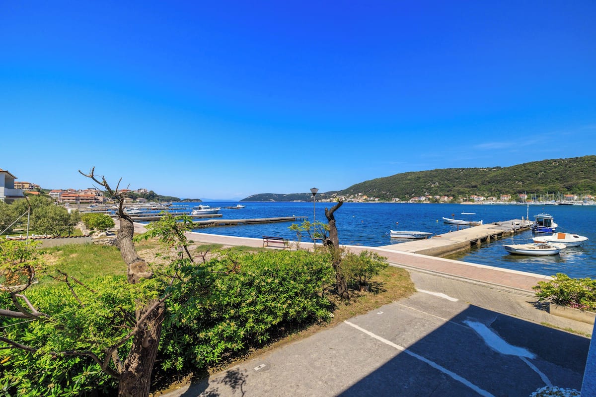 A marina with boats docked, a clear blue sky, and a hilly landscape in the background.