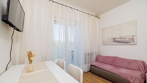 Una stanza con un tavolo bianco, un divano con una coperta rosa, una TV montata a parete e un quadro raffigurante una barca.