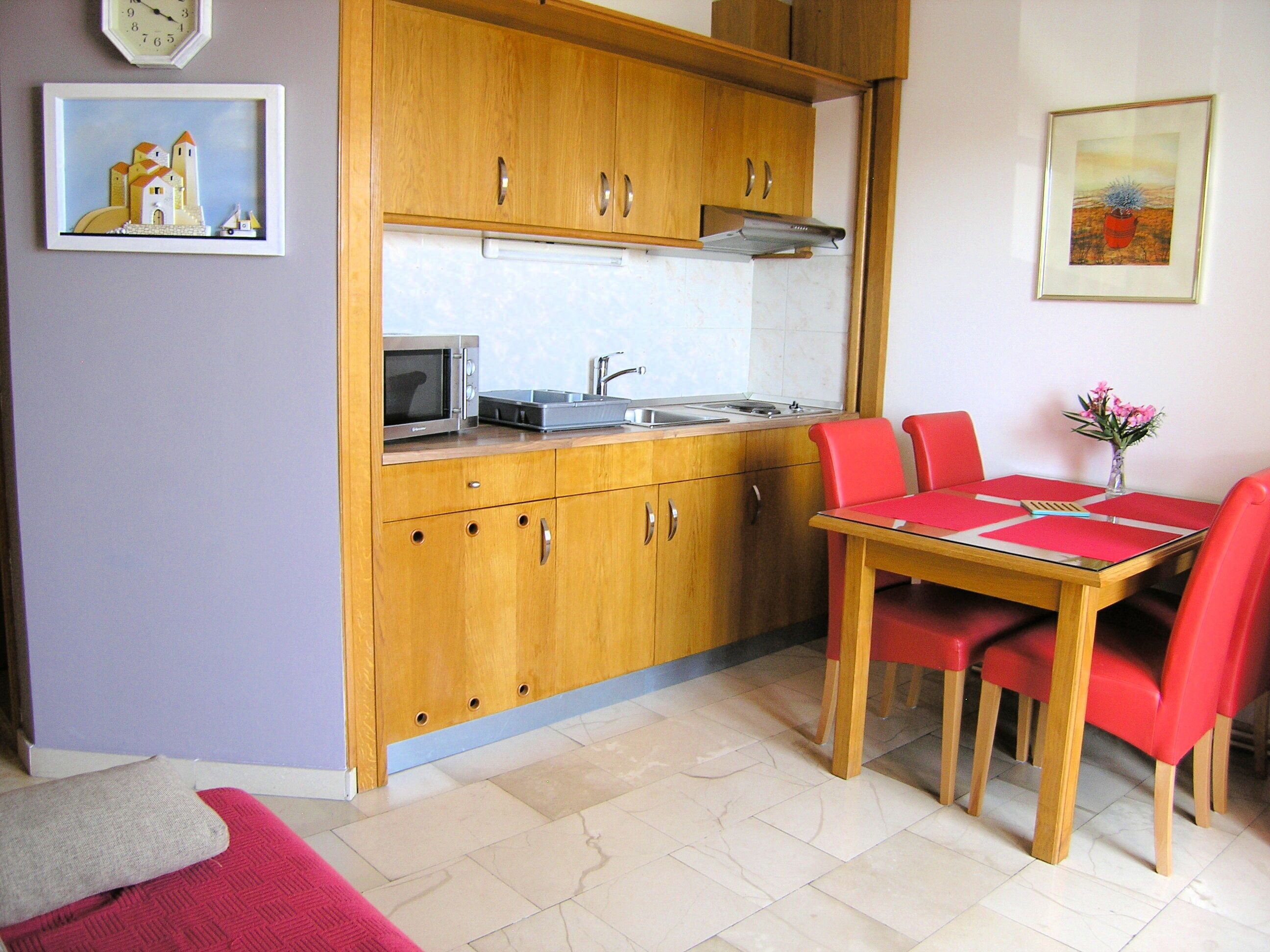 Cocina compacta con armarios de madera, un microondas y un lavabo. Hay una mesa de comedor con sillas rojas y un jarrón con flores.