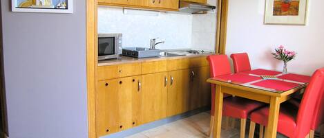 Cocina compacta con armarios de madera, un microondas y un lavabo. Hay una mesa de comedor con sillas rojas y un jarrón con flores.