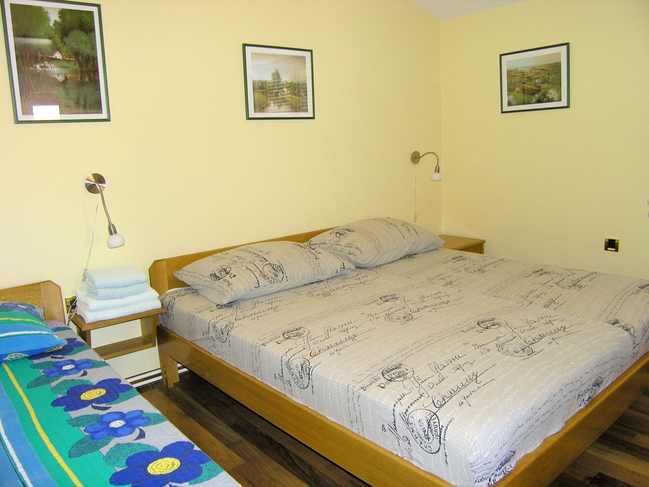 A bedroom with a wooden bed, a bedside table with a lamp, and a wall-mounted light. There are framed pictures on the wall.